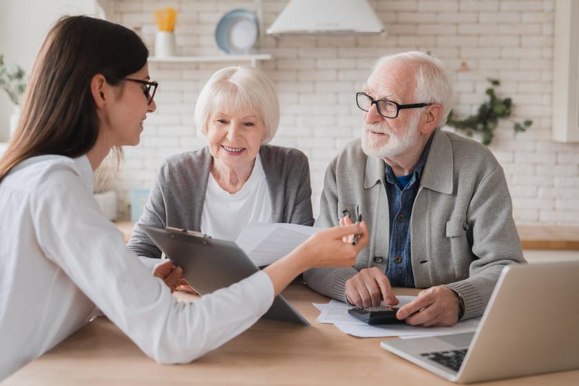 Young woman adviser explains documents to an elderly couple seated at a kitchen table, with a tablet and laptop nearby.