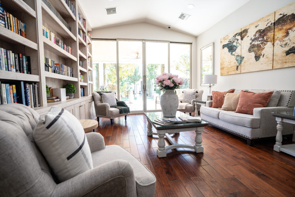 Bright living room with built-in bookshelves along the left wall, gray sofas and armchairs, a glass coffee table, and a three-panel world map artwork above with a vase of pink flowers in the center, sliding glass doors at the back.